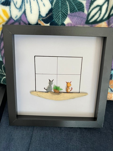 Two pebble cats sitting at a hand-drawn window on a driftwood ledge in a dark 8x8 shadow box frame